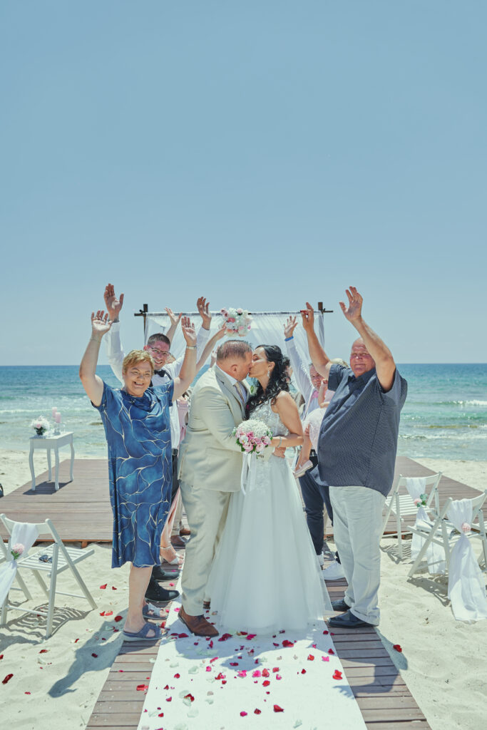 ntimate beach wedding with a few guests seated on white chairs. The couple exchanges vows under a simple floral arch, with the ocean and a clear blue sky in the background. Soft waves lap at the shore, creating a peaceful atmosphere Cyprus