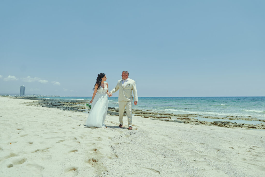 Just married couple posing for a romantic beach photoshoot in Cyprus. The bride wears a flowing white gown, and the groom is in a light suit, standing barefoot on the golden sand. The turquoise sea stretches into the distance, with clear blue skies and a few clouds overhead, creating a dreamy, picturesque backdrop