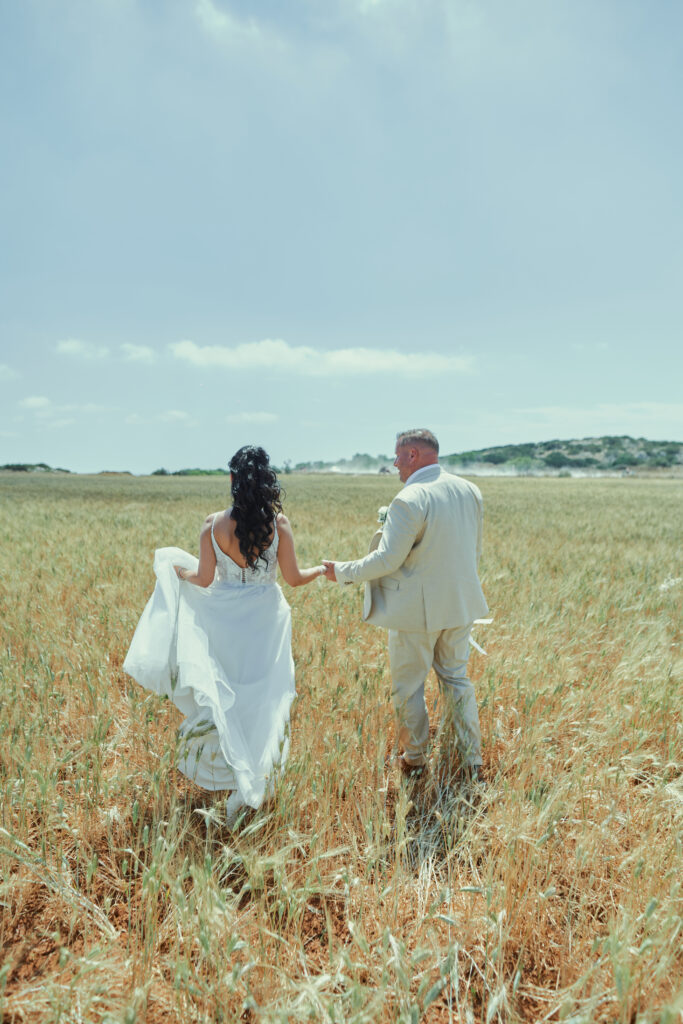 A newlywed couple capturing their love in a beautiful wedding photoshoot in the fields of Cyprus. The bride, wearing a delicate lace gown, and the groom, dressed in a light suit, walk hand-in-hand through a vibrant meadow of wildflowers. The sun sets behind them, casting a warm, golden glow over the lush landscape and the stunning coastline in the distance