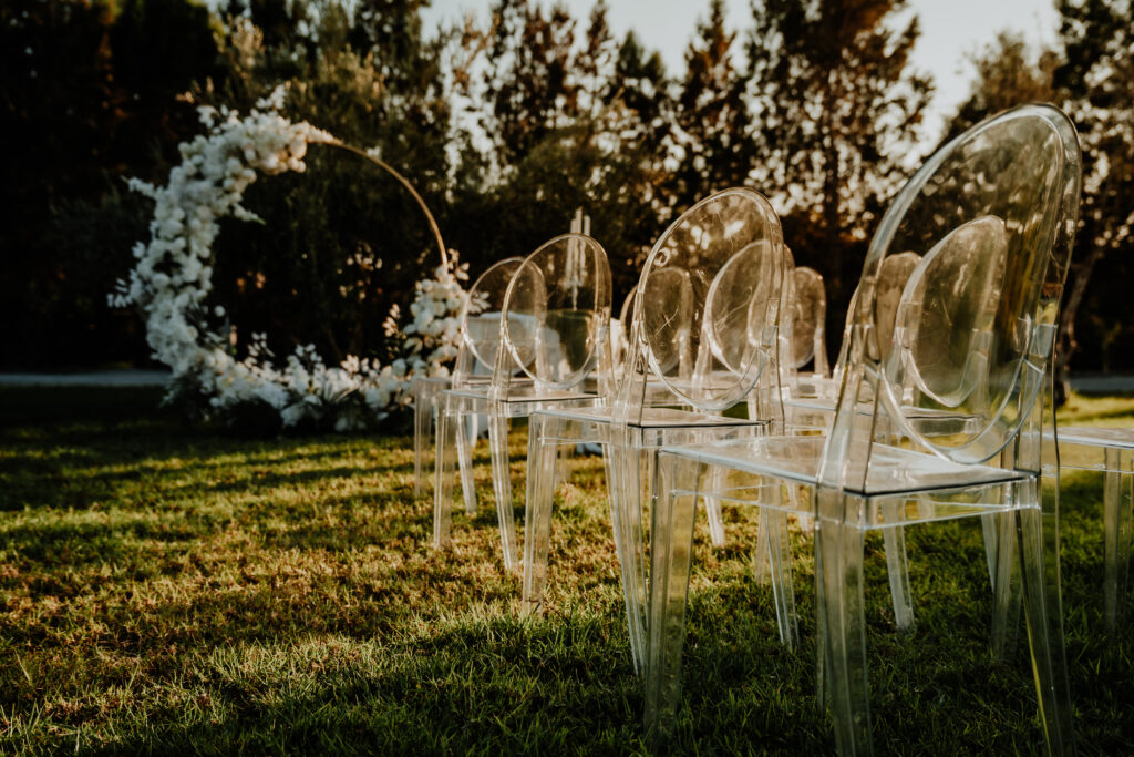 Cyprus garden style wedding ceremony set up including gold round arch with white flowers and clear ceremony chairs