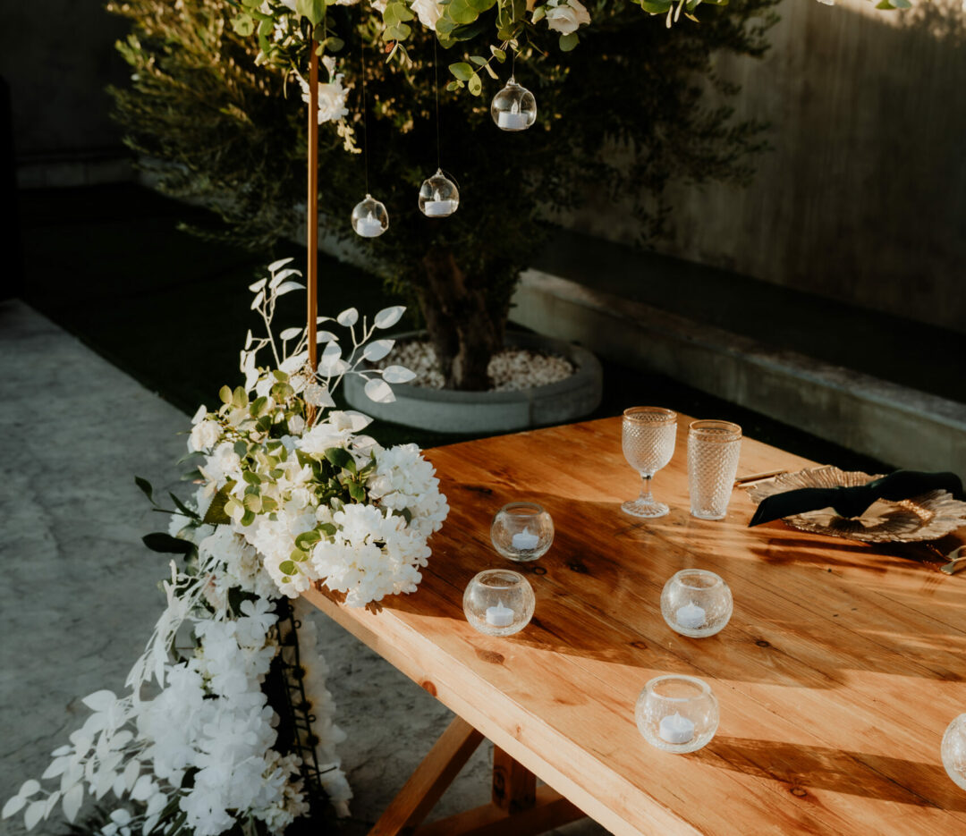 Elegant rustic wedding decor rentals in Cyprus featuring a solid wood harvest table styled with delicate glass tealight globes. The setup includes a modern gold arch adorned with a fresh white floral and eucalyptus runner, creating a sophisticated outdoor ceremony or sweetheart table look for a Mediterranean garden wedding