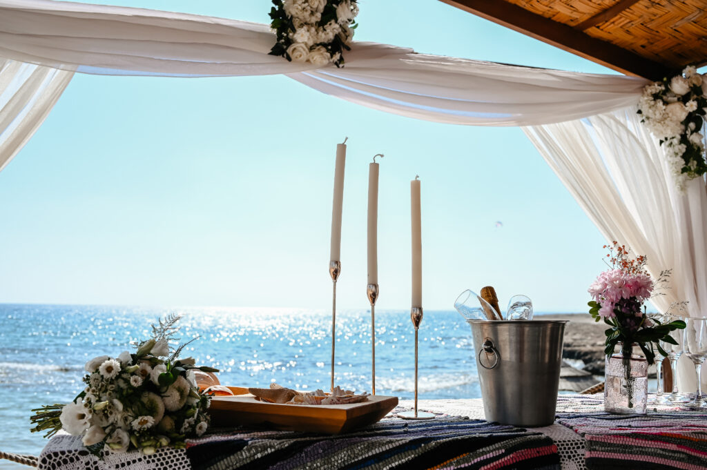 09-09-2024 (1 of 168) Romantic beach wedding setup under a wooden gazebo at Ammos Kampouri, Ayia Napa, featuring white floral arrangements, tall candles, and a stunning ocean backdrop