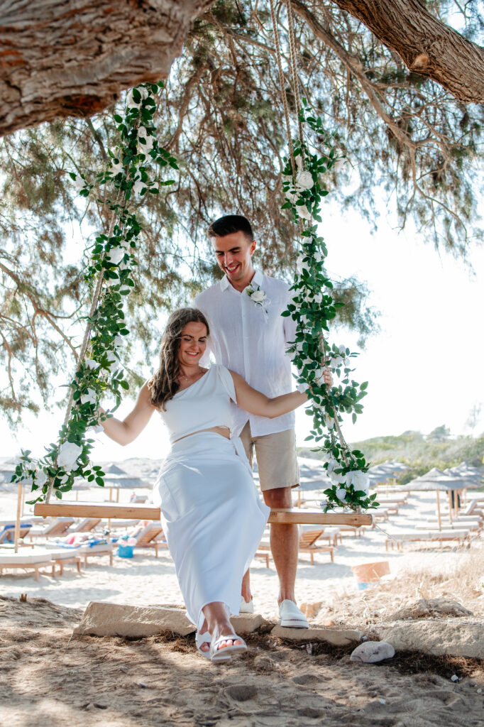 09-09-2024 (152 of 168) Wedding couple joyfully swinging on a swing hanging from an ancient tree at Ammos Kampouri beach, with the serene sea in the background, capturing a playful moment after their ceremony