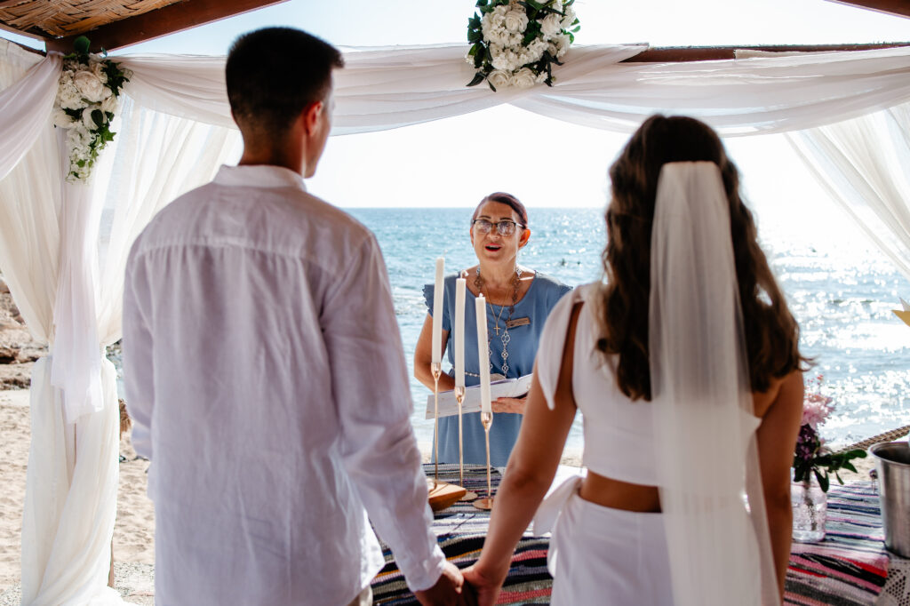 09-09-2024 (29 of 168) Couple holding hands and gazing at the sea while their marriage officiant performs the ceremony under the gazebo at Ammos Kampouri beach, Ayia Napa, Cyprus