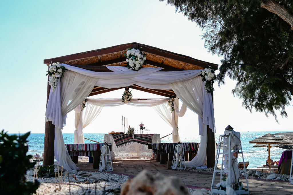 09-09-2024 (8 of 168) Gazebo at Ammos Kampouri beach near Ayia Napa, Cyprus, with white curtains and lanterns draped around it. The wooden seats are covered in colorful fabrics, and the tranquil blue sea stretches out in the background