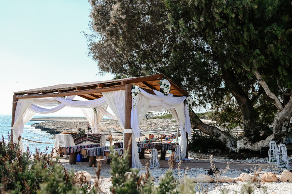 09-09-2024 (9 of 168) Gazebo at Ammos Kampouri beach near Ayia Napa, Cyprus, with white curtains and lanterns draped around it. The wooden seats are covered in colorful fabrics, and the tranquil blue sea stretches out in the background and beautiful tree creates shade during hot summer dates