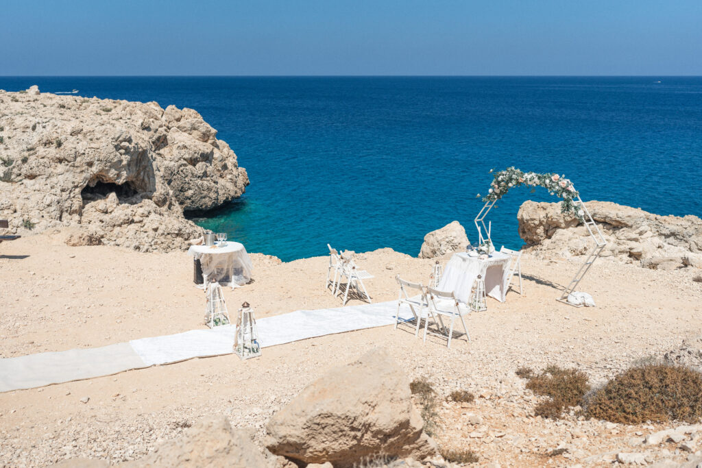 74101192_www.happyimagescyprus.com full view of a wedding decoration set up at Coralli Blue Lagoon Cyprus with a sea backdrop, white ceremony arch with greenery and roses, white carpet and chairs
