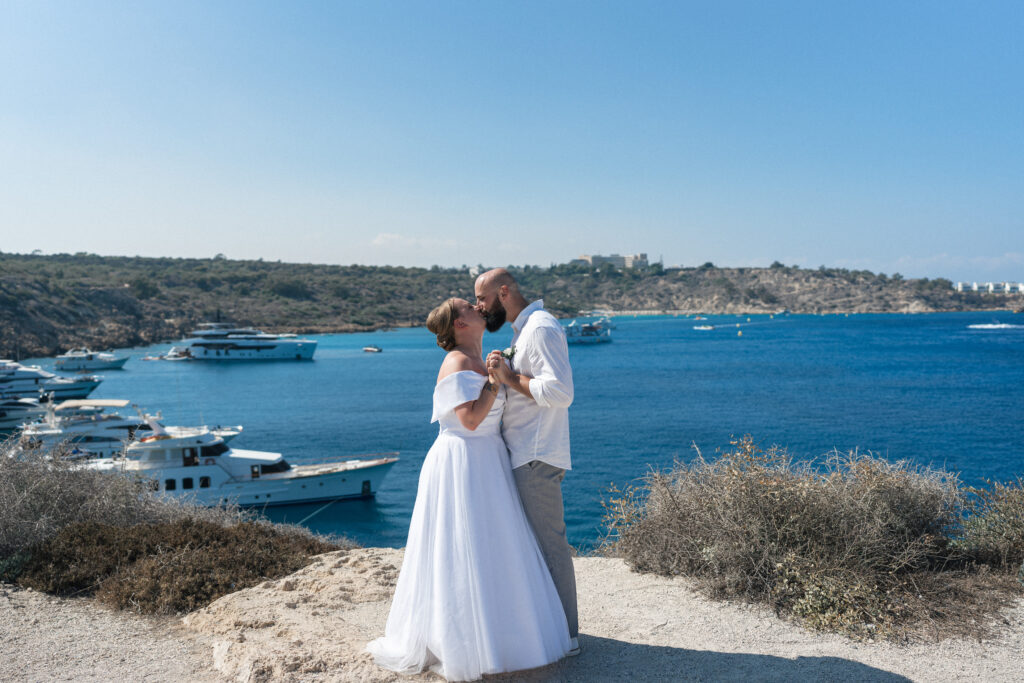 74101386_www.happyimagescyprus.com wedding couple kissing during their photoshoot. There is beautiful sea of Konnos Bay with boats behind them