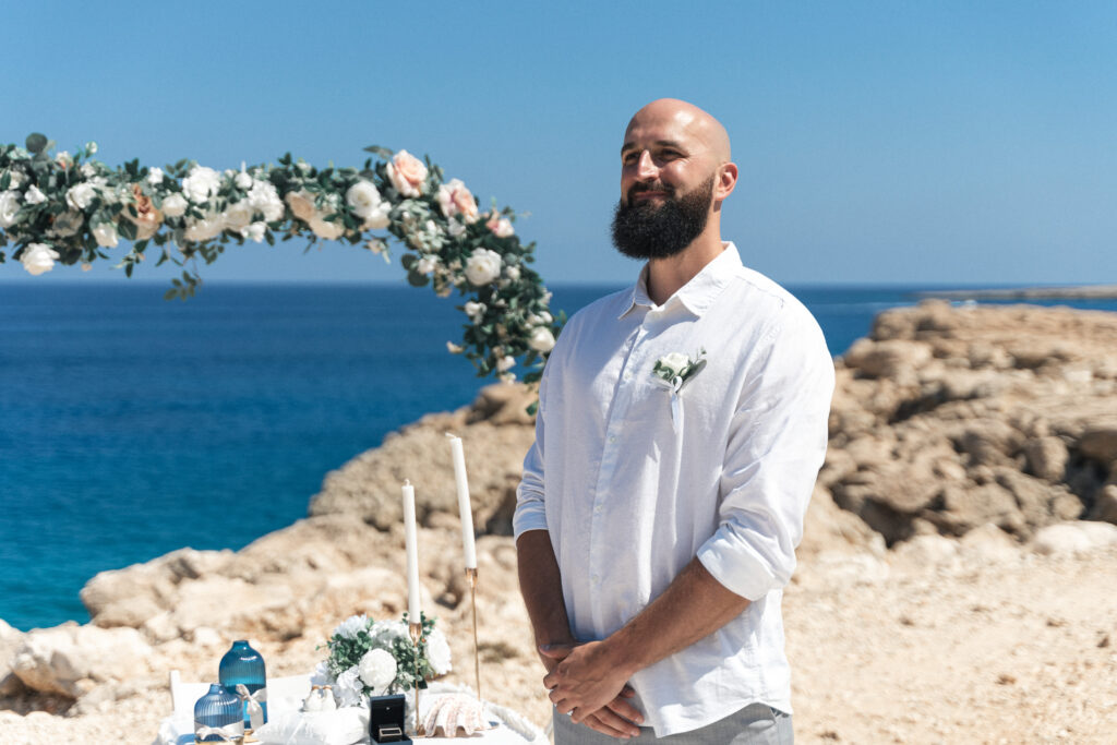 74207507_www.happyimagescyprus.com Groom waiting for a bride in front of floral ceremony arch at Coralli Blue Lagoon near Konnos Bay Cyprus