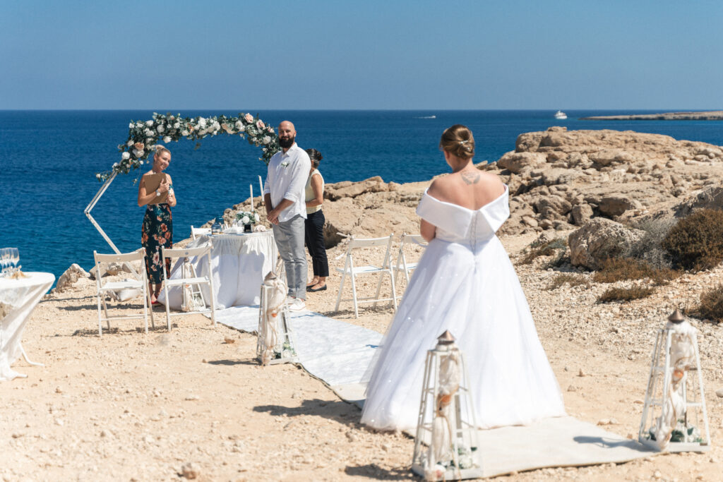 74207602_www.happyimagescyprus.com a beautiful bride walking down the isle as the groom waits for her in front of geometric floral arch at the wedding by the sea in Cyprus
