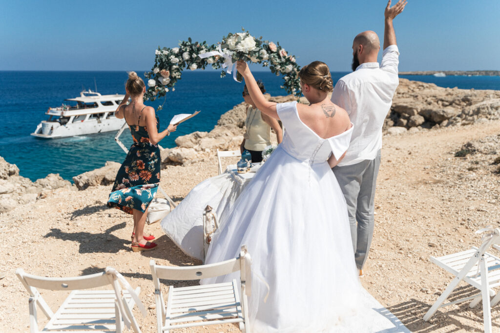 74207722_www.happyimagescyprus.com Wedding couple waving hello to a passing boat at Coralli Blue Lagoon in Cyprus, with the crystal-clear waters and scenic surroundings