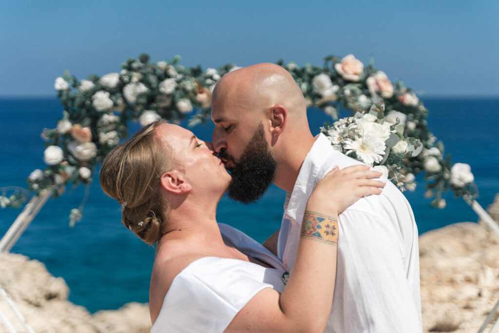 74207767-Edit_www.happyimagescyprus.com magical first kiss of just married couple in front of floral wedding arch and beautiful sea backdrop in Cyprus