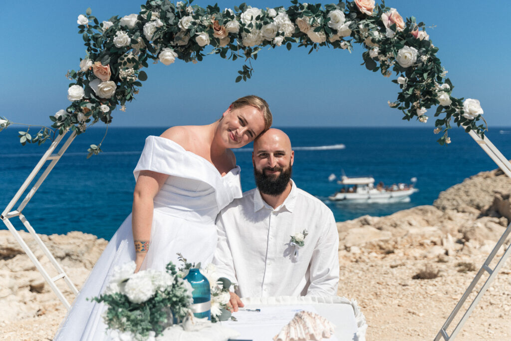 smiling couple in front of white geometric arch with greenery, white and pink flowers signing their marriage certificates during their wedding ceremony in Cyprus