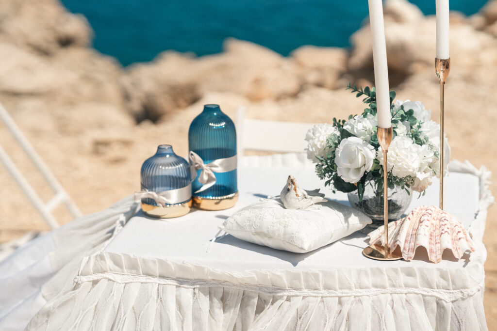 Elegant wedding detail setup at Coralli Blue Lagoon, near Konnos Bay in Cyprus, featuring blue glass decorations, white florals, candles, and a shell accent by the sea.