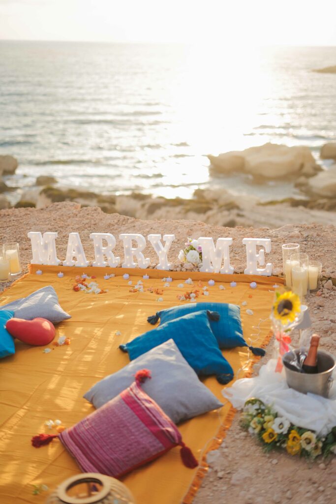 Close-up of a romantic beachside proposal setup in Cyprus, featuring a champagne bottle in an ice bucket, two glasses, a sunflower, and cozy decor with flowers and candles.