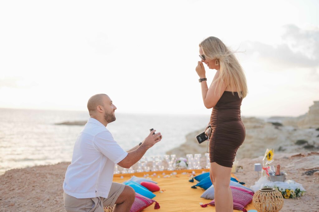 Romantic beachside proposal in Cyprus with a man on one knee presenting a ring to his partner, set against a stunning coastal sunset with a beautifully arranged picnic setup.