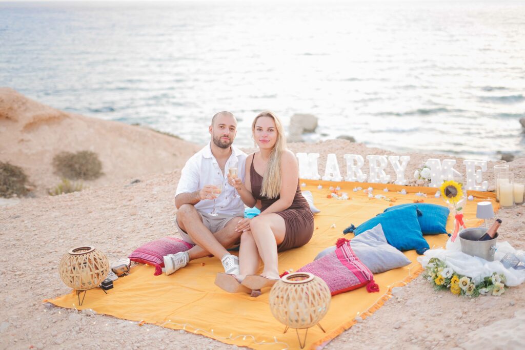 happy couple sitting on a yellow blanket in front of Marry me sign enjoying their marriage proposal in Cyprus