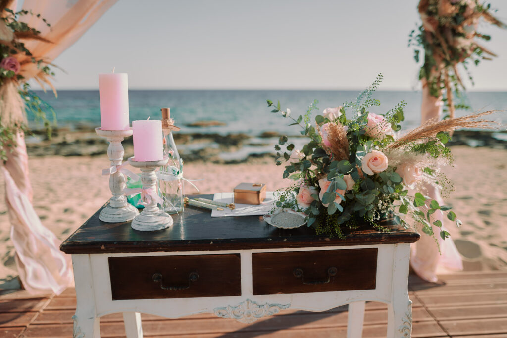 Romantic beach wedding ceremony setup in Ayia Thekla, Cyprus, featuring a vintage wooden signing table decorated with pastel pink candles, floral arrangements, and wedding accessories. Elegant wedding decoration hire and rental available for dreamy seaside weddings