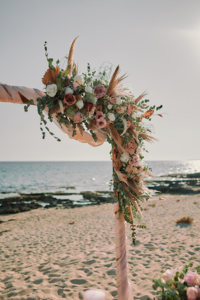 Elegant beach rustic wooden wedding arch in Cyprus, beautifully decorated with blush and white roses, eucalyptus, pampas grass, and soft draping fabric. Perfect wedding decoration hire and rental for a romantic seaside ceremony.