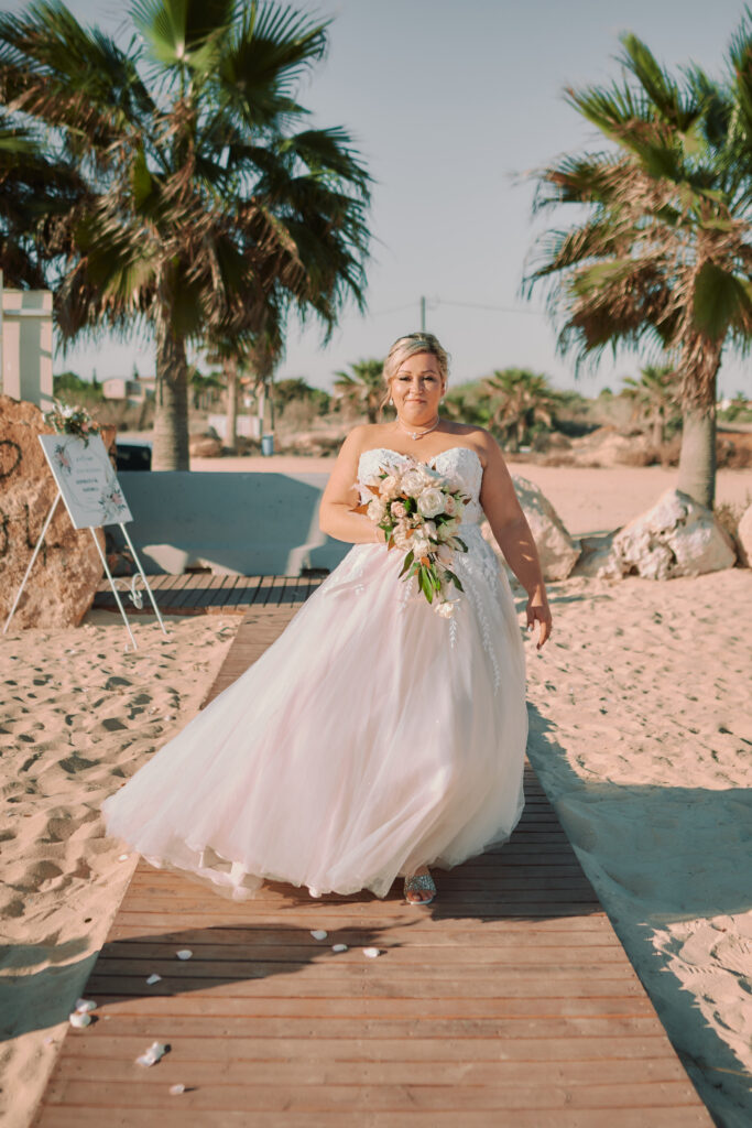 beautiful bride walking down the wooden deck at Sirens beach venue in Cyprus holding rose flower bouquet with palm trees behind her