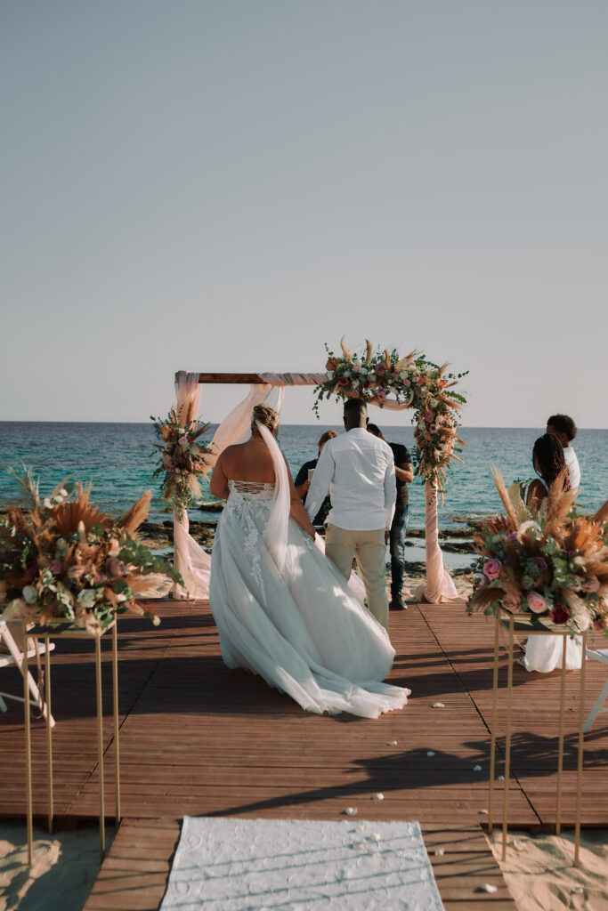 beautiful professional wedding photography of bride and groom in front of wooden rustic ceremony arch with roses, greenery and pampas grass at Sirens beach venue in Ayia Napa Cyprus. One of the best places for a ceremony in Greek islands