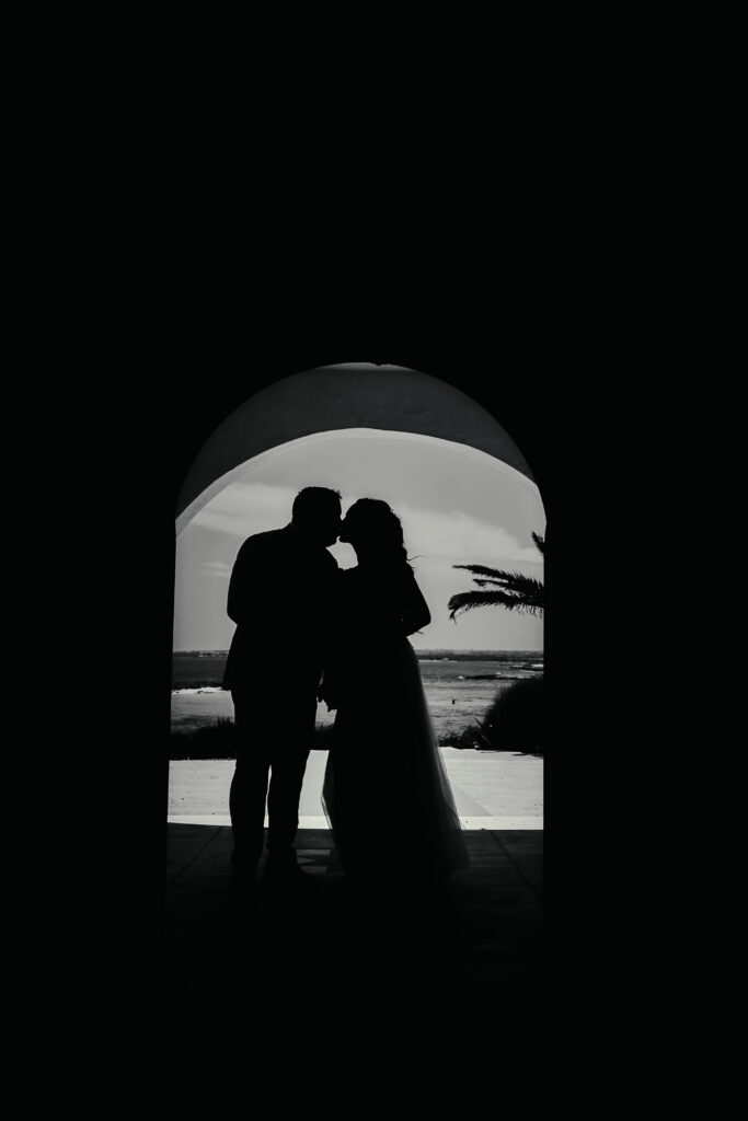 Romantic silhouette of a bride and groom kissing under a stone archway at their beach wedding in Ayia Napa, Cyprus, with the sea and palm trees in the background.