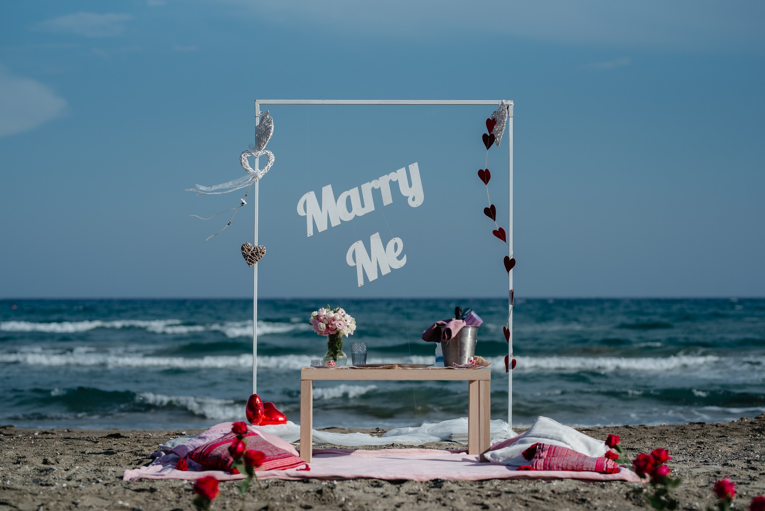 romantic beach picnic marriage proposal in Larnaca Cyprus, featuring white marry me sign, pillows, wooden table and fresh flowers