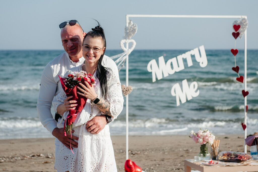Romantic beach marriage proposal in white colors, hanging Marry me sign and heart decorations