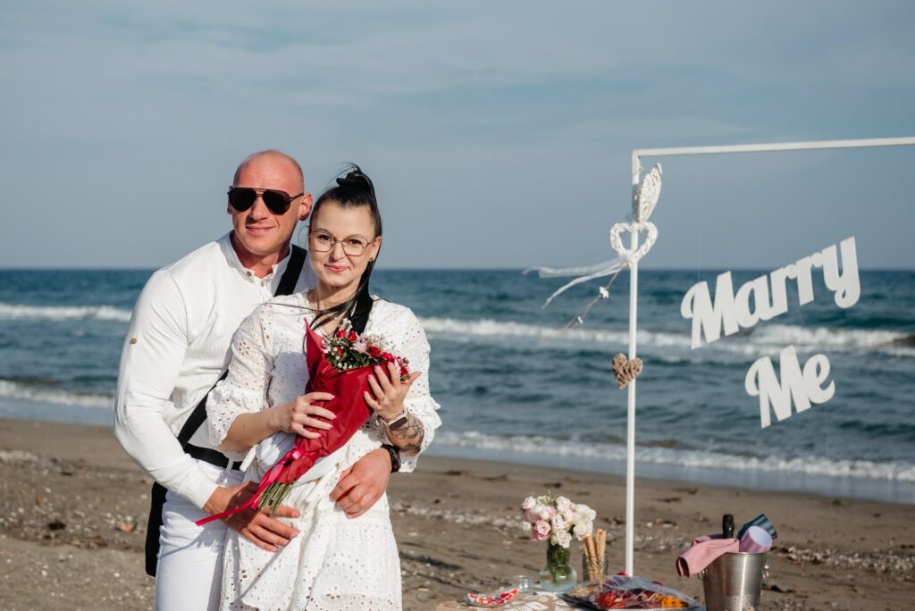 Cyprus beach wedding proposal couple standing in front of white Marry me sign and picnic table during their beach marriage proposal