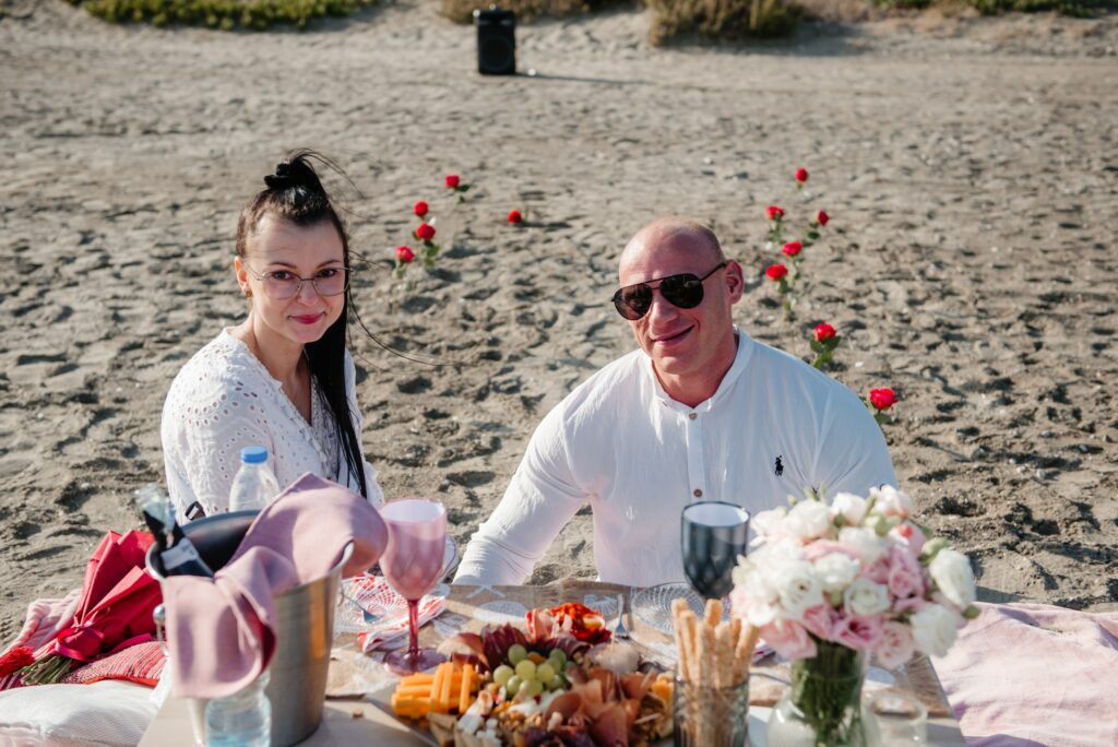 newly engaged couple is enjoying their romantic proposal picnic with meat and cheese platter, Prosecco and floral decor at the beach near Larnaca