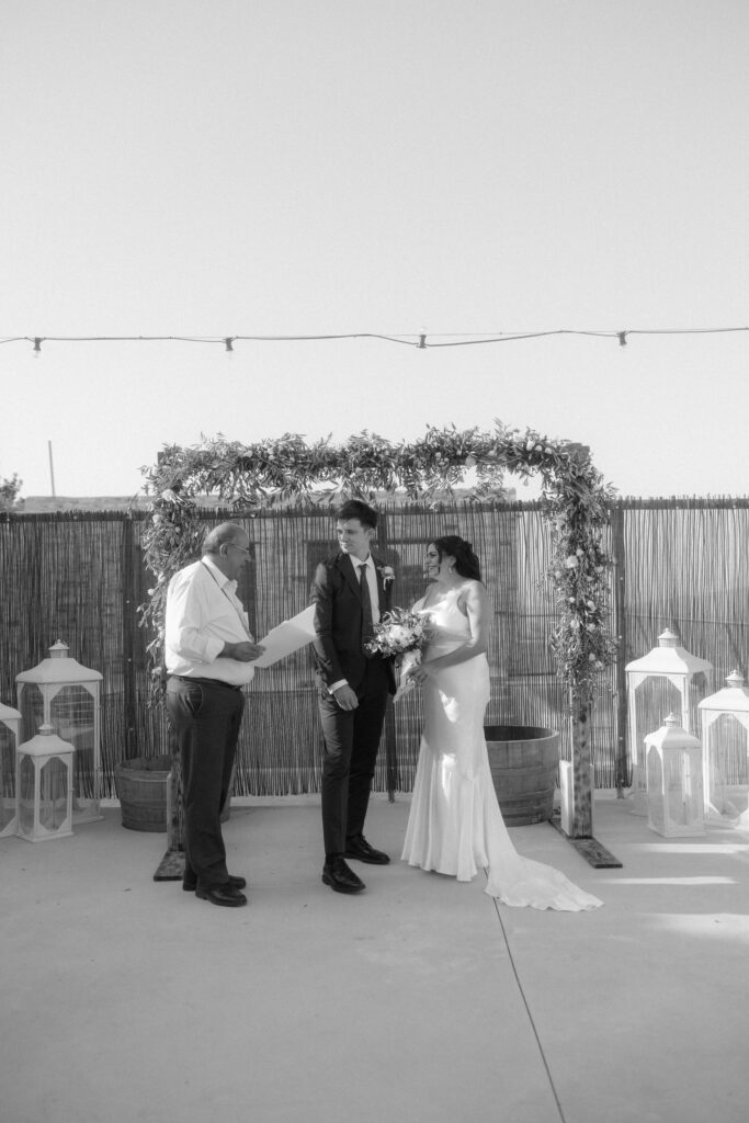black and white wedding photography. Bride and groom standing in front of their rustic floral ceremony arch in Cyprus