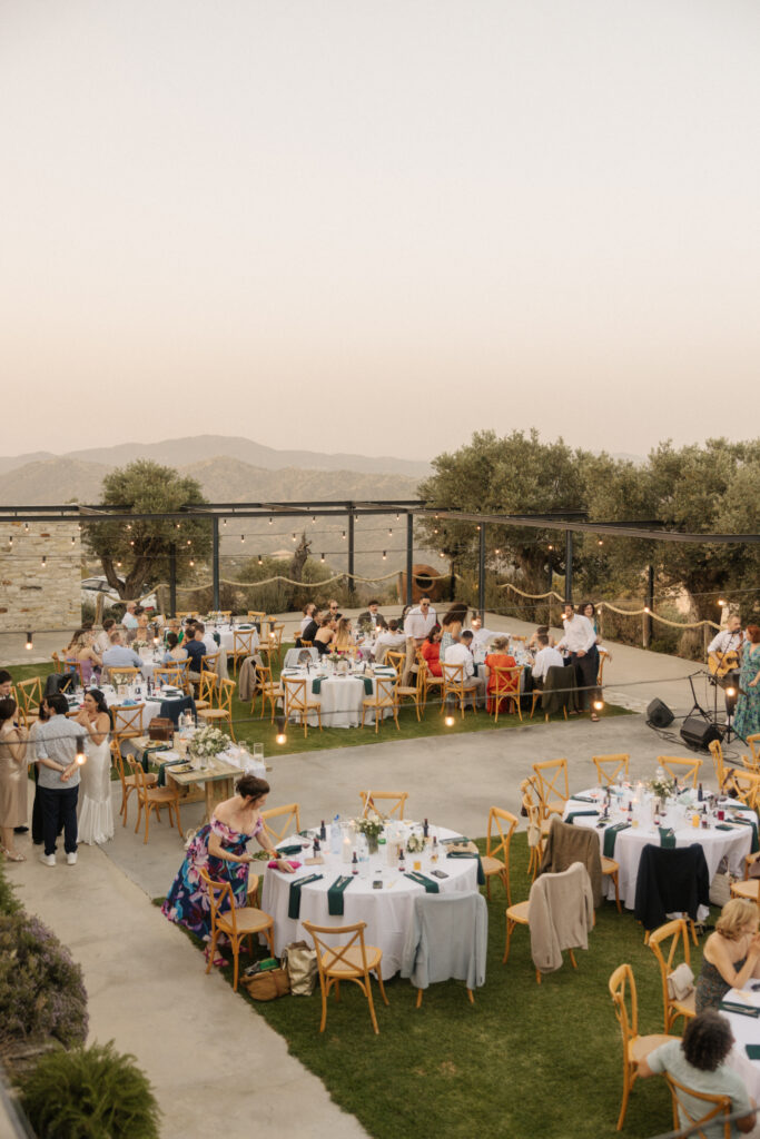 Chic outdoor wedding reception at Dafermou Winery in Cyprus with scenic mountain views, string lights, wooden cross-back chairs, and round tables decorated with green napkins and floral centerpieces from decoration rental services