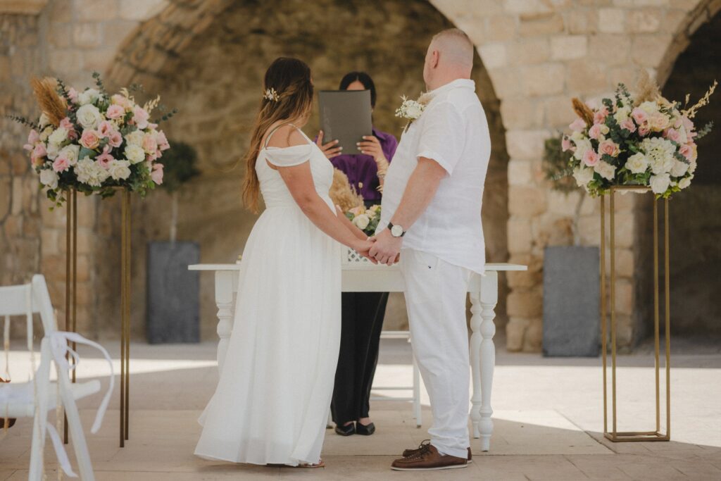 Gieroskipou town hall gardens rustic wedding ceremony A bride and groom holding hands during an intimate civil ceremony at Gieroskipou Town Hall, featuring a rustic stone archway, elegant floral arrangements on gold stands, and a white ceremonial table.