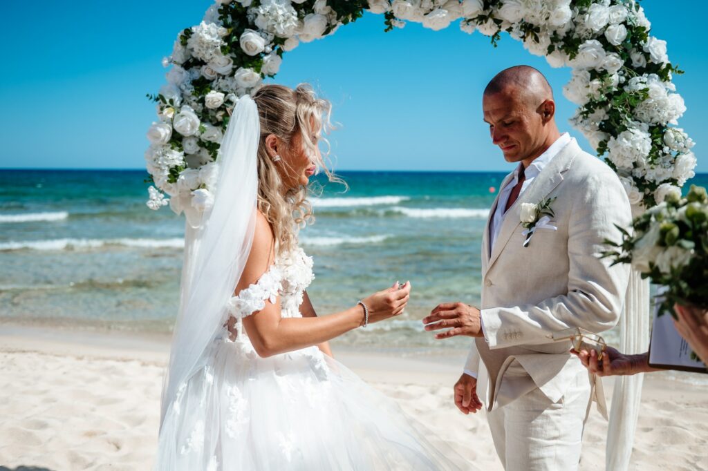 Bride and groom exchanging wedding rings in front of white and green floral arch during beach ceremony in Cyprus