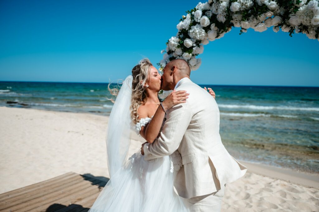 Bride and groom sharing their first kiss in front of white and green floral arch during beach ceremony at Sirens Beach in Cyprus