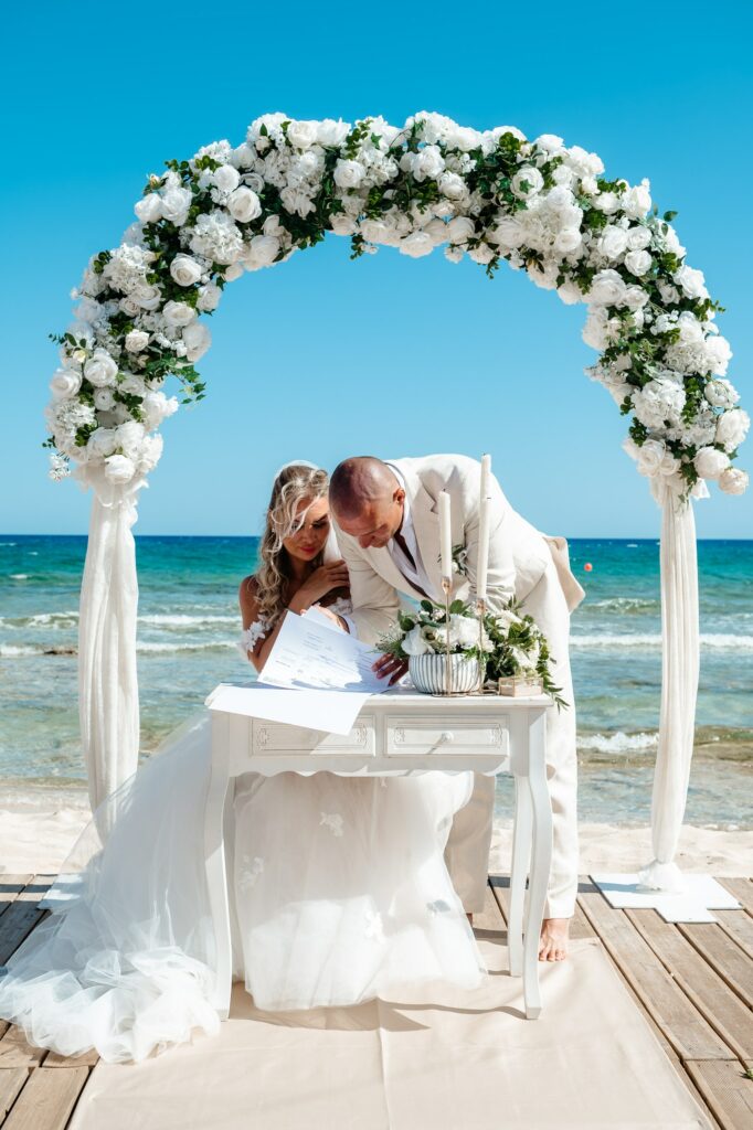 Bride and groom signing marriage certificate on white rustic table in front of white and green round floral arch with Cyprus sea backdrop at Sirens Beach