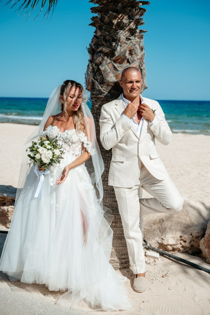 Bride and groom posing by a palm tree during a beach wedding at Sirens Beach in Cyprus, bride holding white rose bouquet and groom wearing beige suit