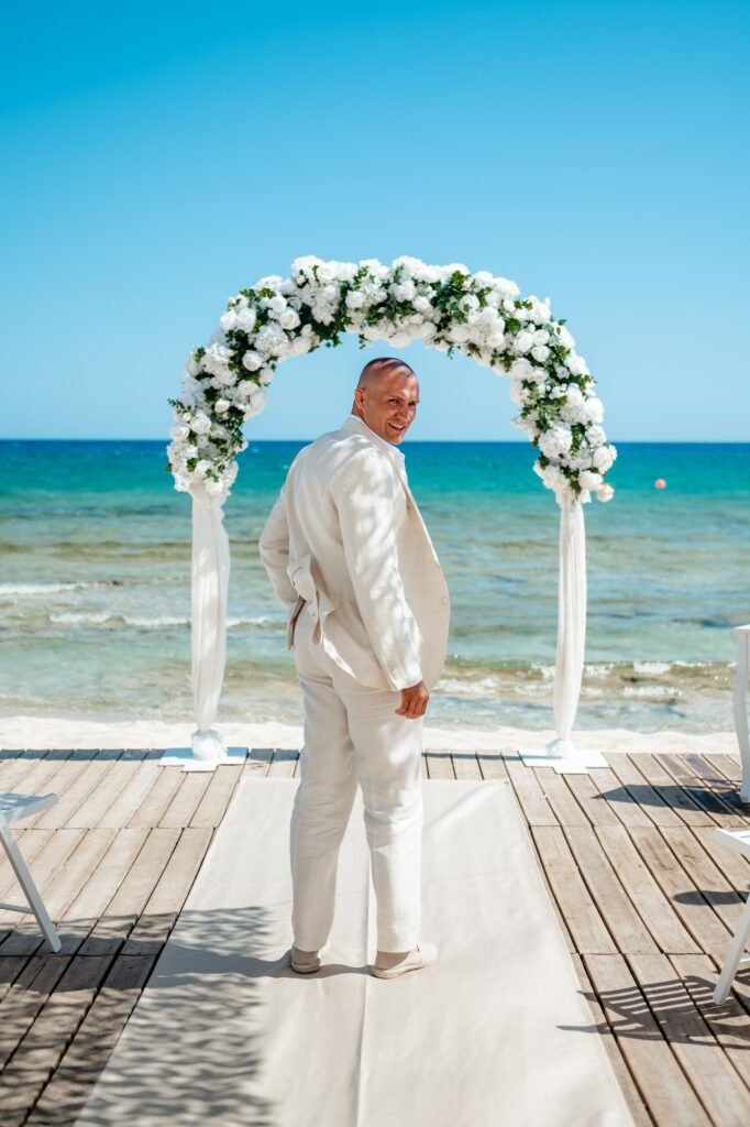 Groom in beige suit standing in front of white and green wedding ceremony arch at a beach in Cyprus