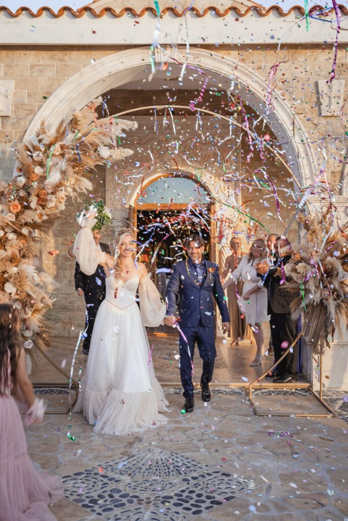 Newlywed couple walking out of St. Epifanios Church in Ayia Napa, Cyprus, surrounded by guests as colorful confetti fireworks fall in the air, with pampas grass, dried floral wedding decorations, and stone archway entrance in the background.