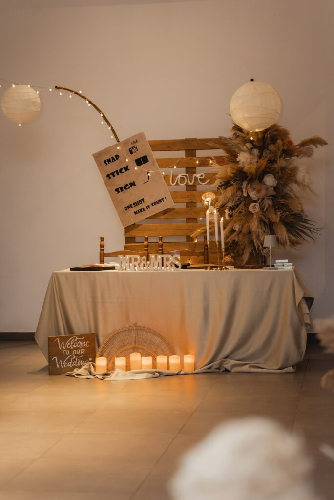 Boho wedding welcome table setup with beige tablecloth, wooden pallet backdrop, pampas grass and dried floral arrangement, glowing candles, paper lanterns, string lights, ‘Mr & Mrs’ sign, and wedding décor details in warm neutral tones