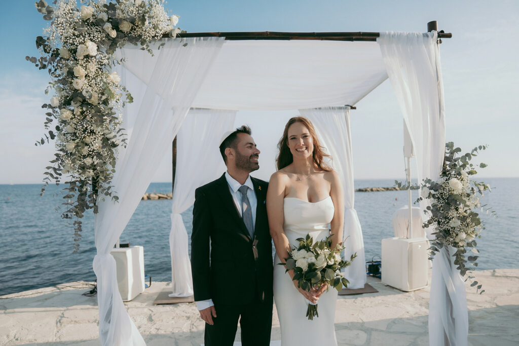 Bride and groom standing under a floral chuppah by the sea at the Four Seasons Hotel Limassol, Cyprus, during an elegant beachfront wedding ceremony.