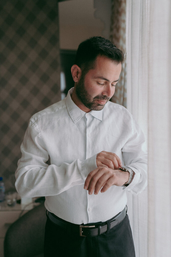 Groom getting ready for the ceremony in hotel room fixing his white shirt