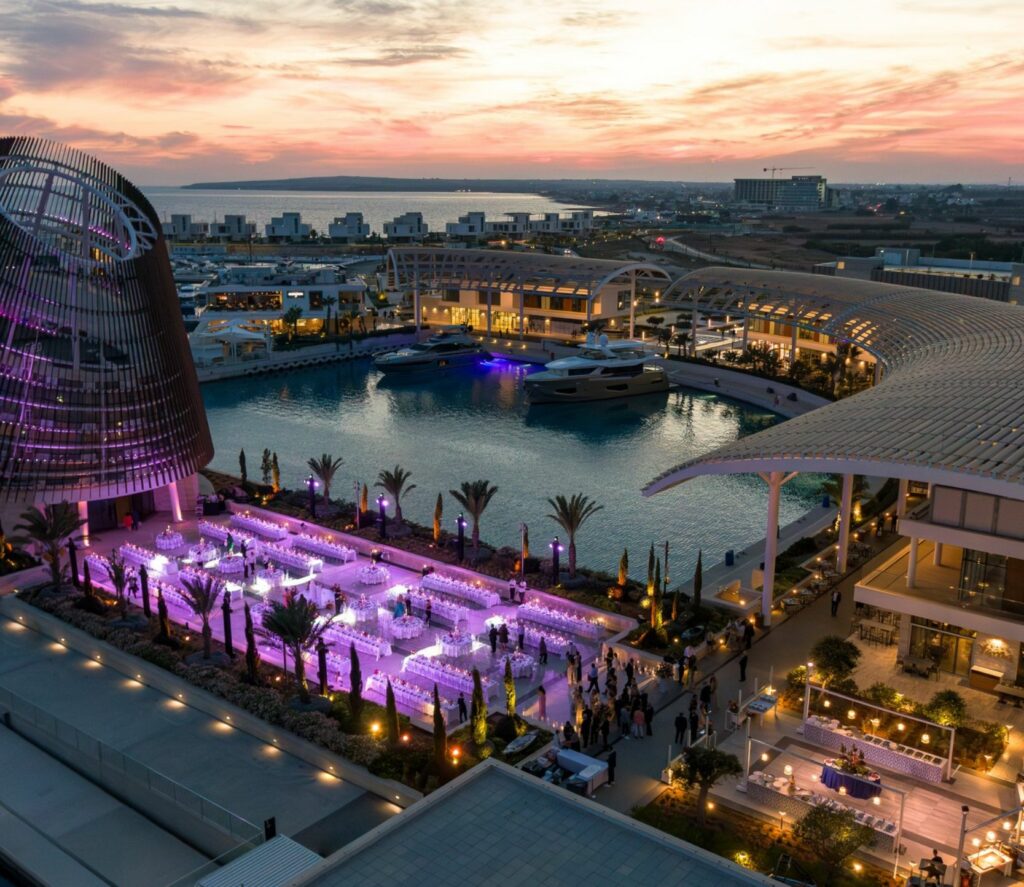 Ultra-modern wedding reception at Ayia Napa Marina, Cyprus, featuring an expansive outdoor setup with purple ambient uplighting and white Napoleon chairs. The luxury event styling overlooks the harbor at sunset, showcasing a high-end gala dinner with sophisticated floral centerpieces and professional event production.