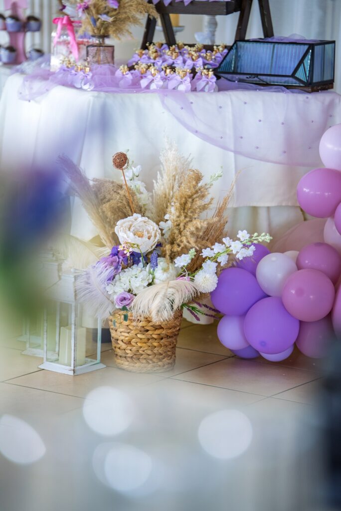 A purple-themed baptism candy table in Cyprus with a pampas grass floor arrangement in a wicker basket, lilac balloons, and delicate tulle table draping.