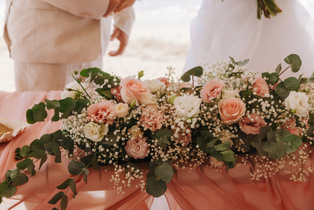 Detailed view of a romantic wedding table centerpiece featuring a lush arrangement of peach roses, white eustoma, and eucalyptus greenery. This ceremony table decor in Cyprus includes soft blush pink chiffon draping, perfect for a dreamy beachfront or garden wedding looking for premium floral and linen rentals.