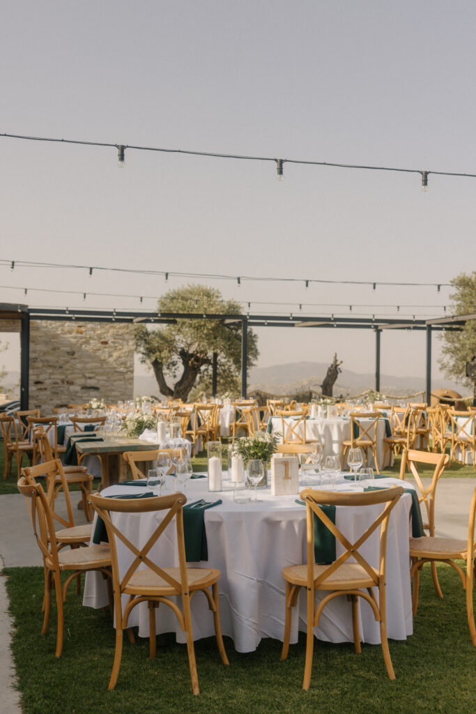 Rustic winery wedding decoration in Cyprus featuring wooden crossback chairs and deep forest green napkins. The outdoor reception setup at a stone estate includes bistro festoon lighting, minimalist white floral arrangements, and scenic mountain views for an authentic Mediterranean winery celebration.