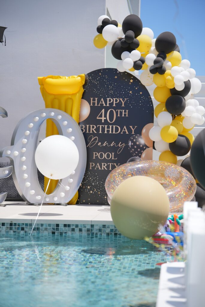 40th birthday pool party rentals in Cyprus featuring a black custom backdrop, lighted marquee numbers, and a black, white, and yellow balloon arch by a villa pool.