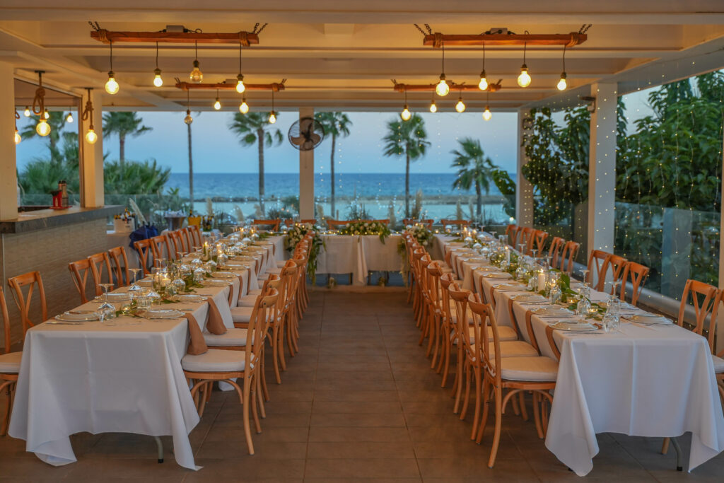 Elegant wedding reception at Nissaki venue in Ayia Napa, featuring long banquet tables with white linens and wooden crossback chairs. The coastal decor includes lush greenery runners, warm Edison bulb overhead lighting, and a stunning Mediterranean sea view, perfect for a modern beachfront wedding in Cyprus.