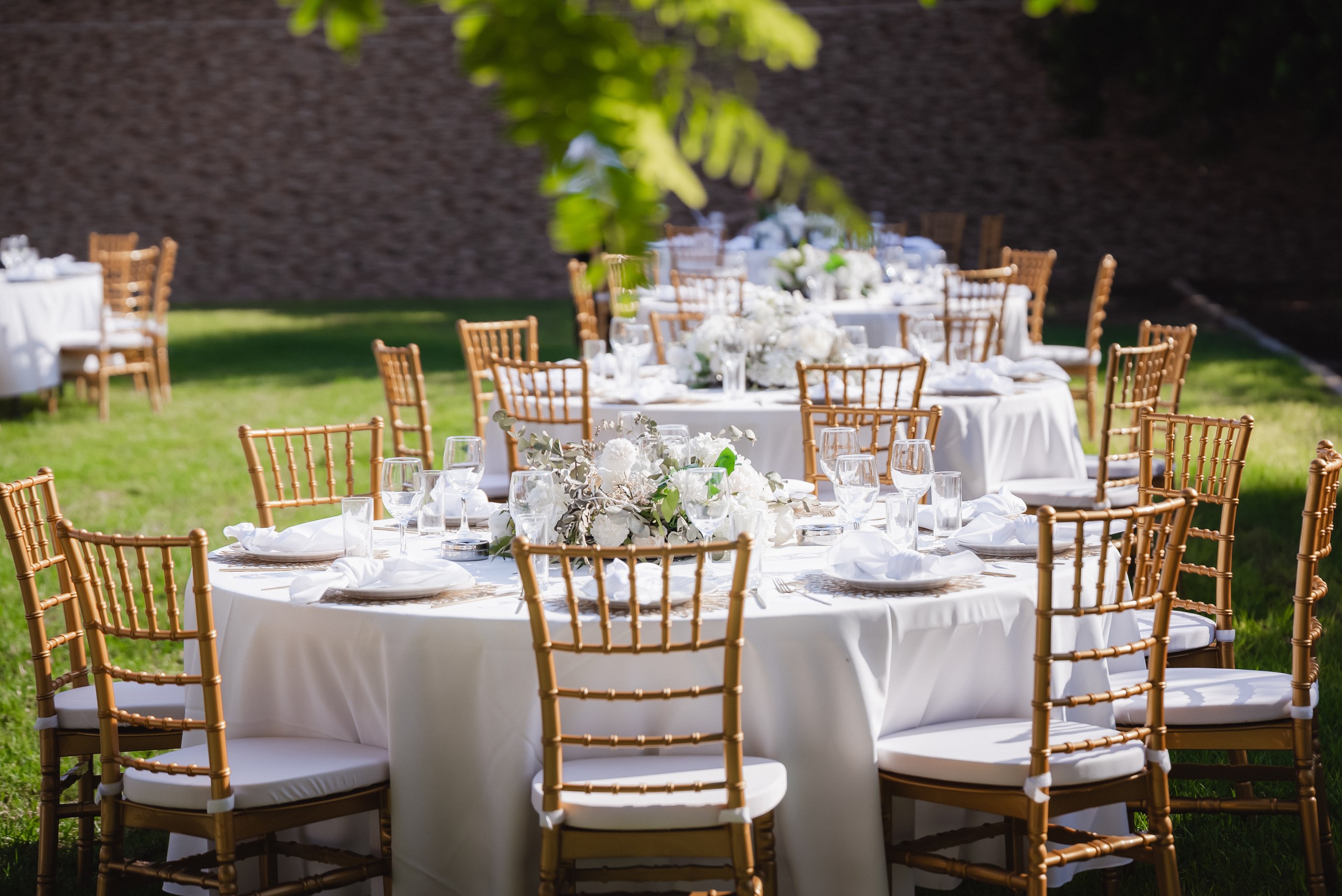 Gold Chiavari chairs and crisp white linen tables with lush white floral centerpieces for an outdoor wedding reception in Cyprus. This scenic setup features luxury wedding styling for a destination wedding, including elegant glassware and garden venue decor.
