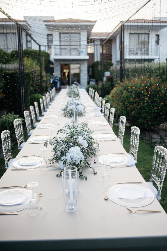 Elegant private villa wedding in Cyprus featuring a long banquet table under a stunning fairy light canopy. This garden reception setup includes clear Napoleon ghost chairs, gold cutlery, and fresh flower arrangements of white hydrangeas and olive leaves, showcasing premium wedding decoration rentals for an intimate estate celebration.